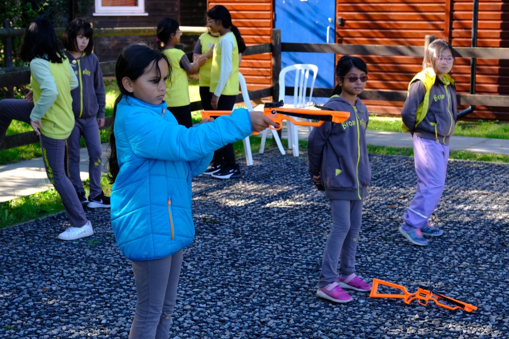 A girl in a blue coat holds a yellow plastic suction-cup crossbow as she fires it. Two other girls wait for their turns.