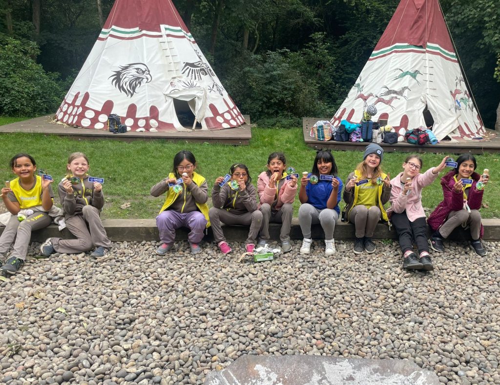 A line of Brownies sitting in front of two Teepees. They're eating ice creams and holding up two fabric badges.