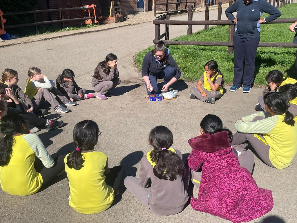 Brownies in their uniforms sitting on the ground in the sunshine looking at their leader who has an open tin and is showing them flint and steel.