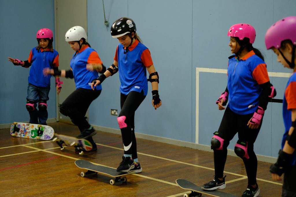 Three Guides in uniform wearing safety helmets, gloves, and elbow and knee pads. In a hall, they rest a foot on the end of their skateboards.
