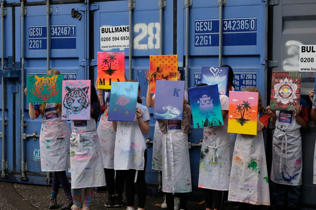 A group of girls in aprons standing and holding their colourful paintings up in front of their faces.