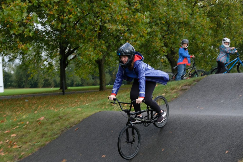 A Guide cycles over a hump on the tree-lined track, her blue coat flying out behind her as she goes. Another girl waits with her bike in the background.