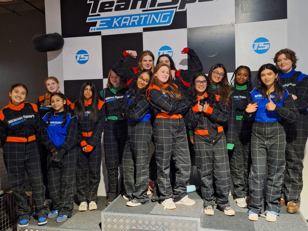 A group of girls in their karting jumpsuits standing on and around the winners' podium. Two in the centre stand with their arms folded and with their backs to each other. Two behind flex their muscles.