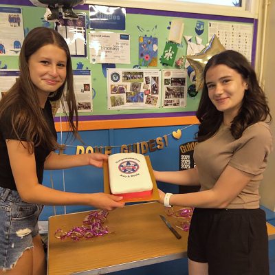 Two girls with long dark hair smiling and holding a rectangular cake with "Congratulations" above a star image.