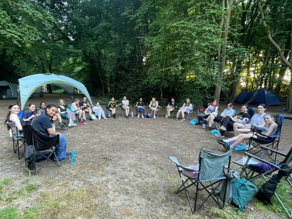 A group of girls sitting on camp chairs in a circle with a tent in the background.